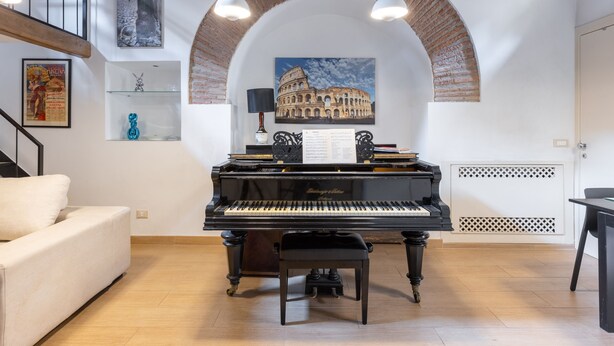 A living room with a grand piano, a white sofa, a staircase, and a large arched brick wall.