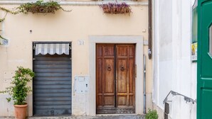 A narrow street with a building featuring a closed shutter door, a wooden double door, and a balcony with potted plants.