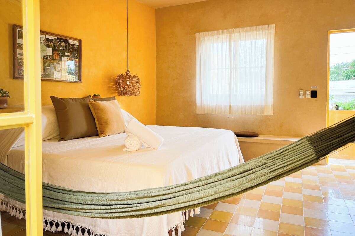A bedroom with a bed, a hammock, a window with curtains, and a wall-mounted light fixture.