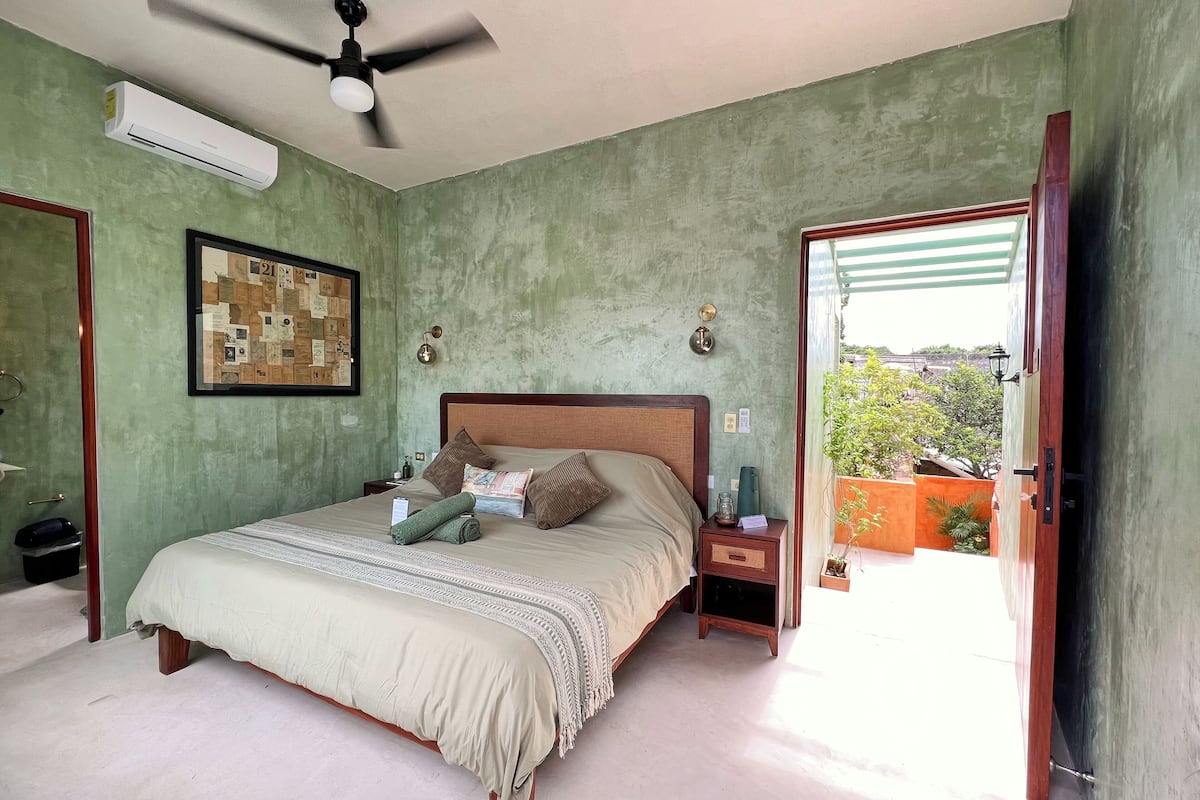 A bedroom with a bed, a ceiling fan, an air conditioning unit, a nightstand, a framed picture, and a view of the outside.