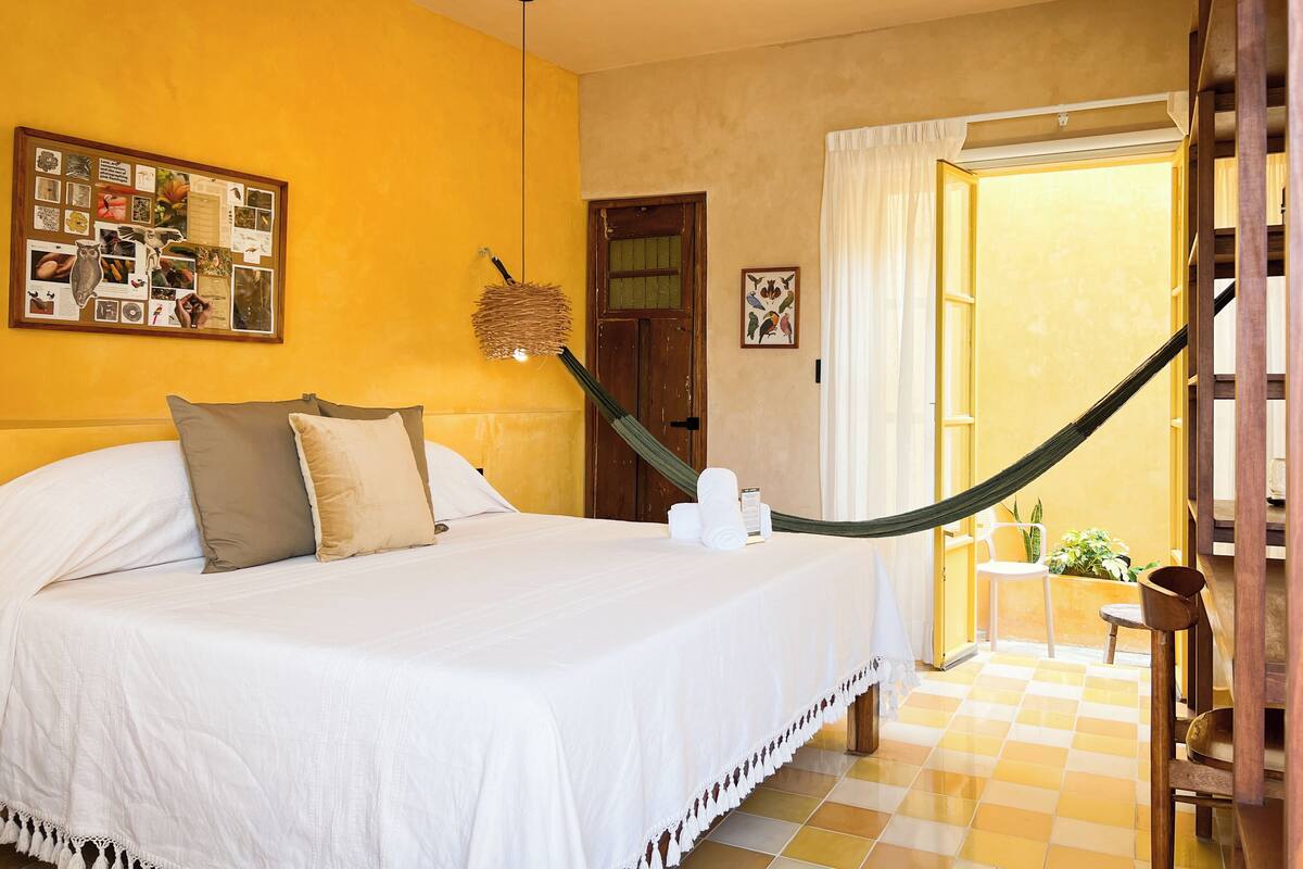 A hotel room with a bed, a bulletin board, a hammock, and a view of the outside.
