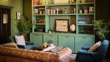 A cozy living room with a brown leather sofa, teal armchairs, and a green bookshelf filled with books and decorative items.