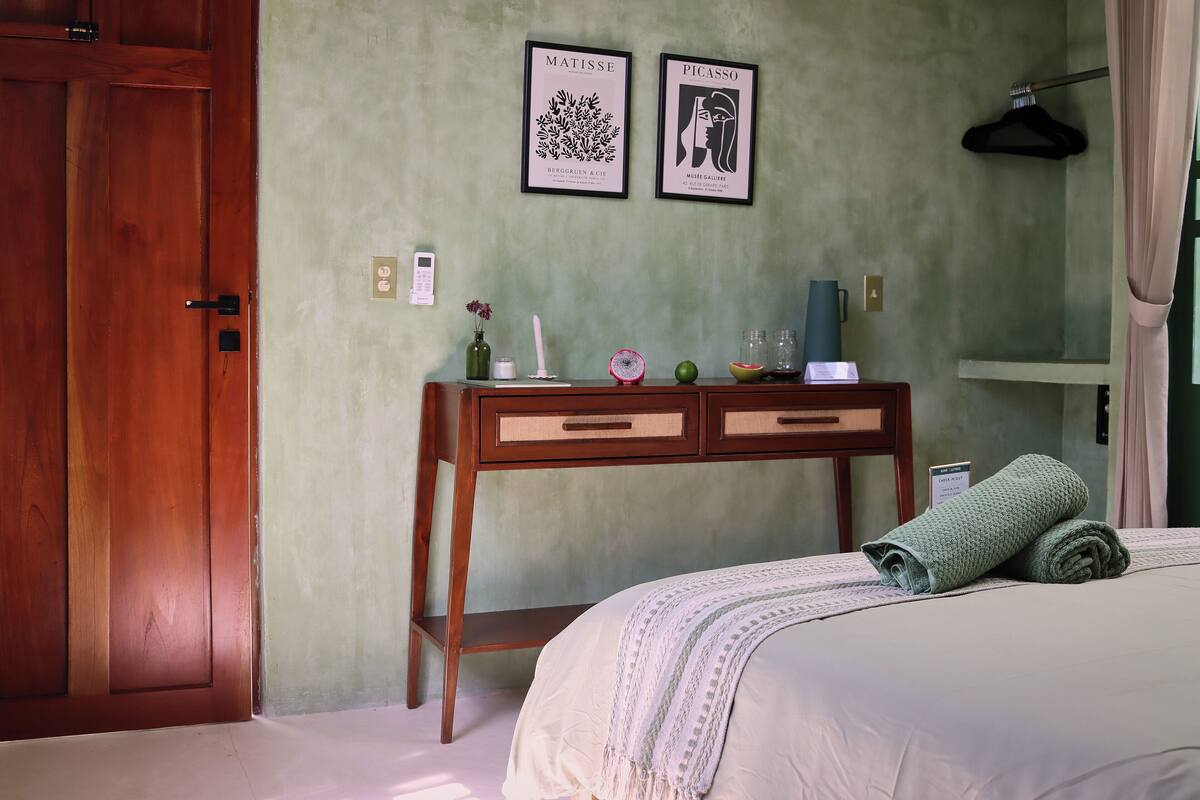 A hotel room with a wooden door, a bed with a rolled green towel, a bedside table with a vase, a candle, and a framed artwork of Matisse and Picasso.