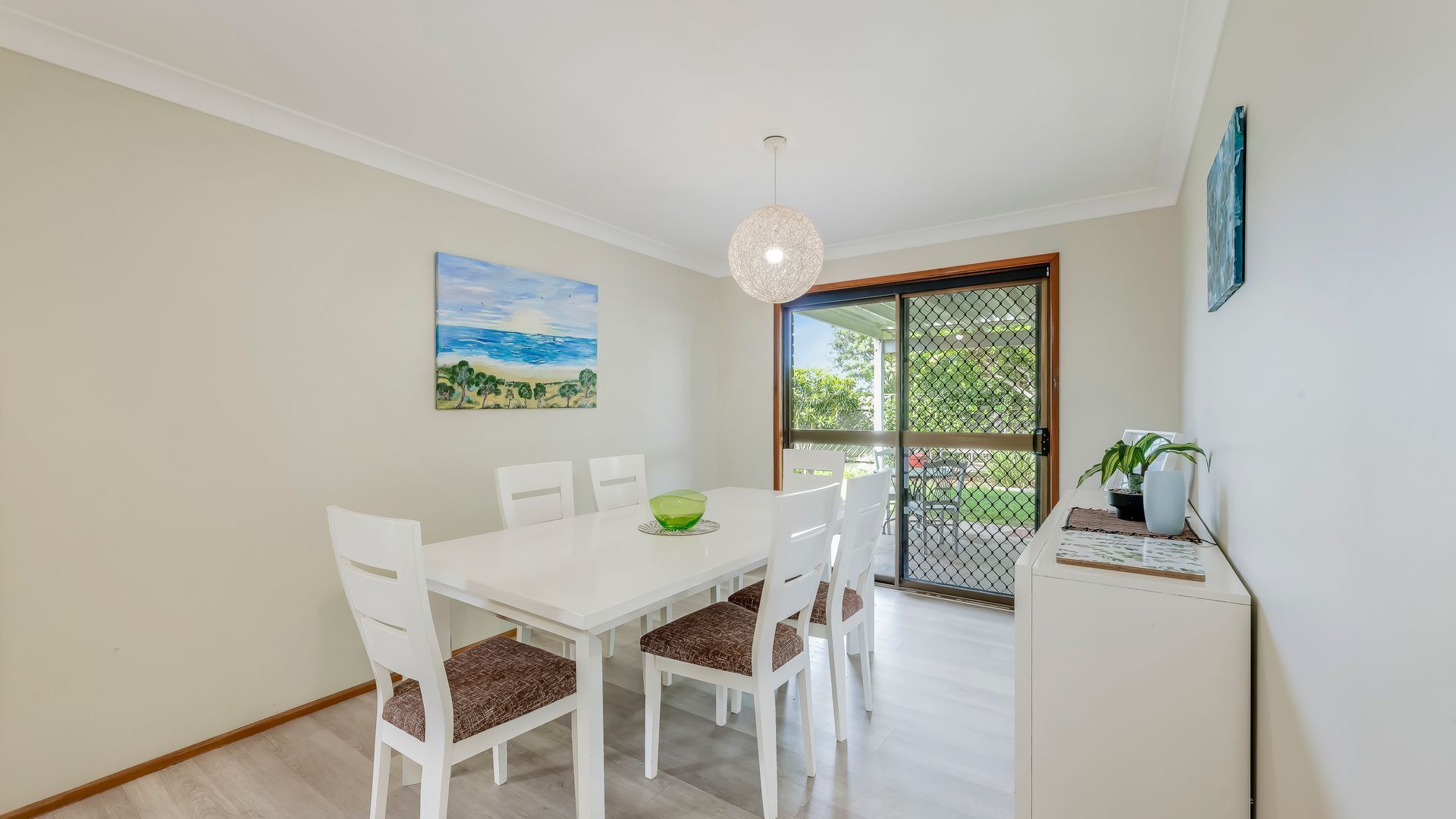 A dining area with a white table and chairs, a green bowl, and a painting of a landscape.