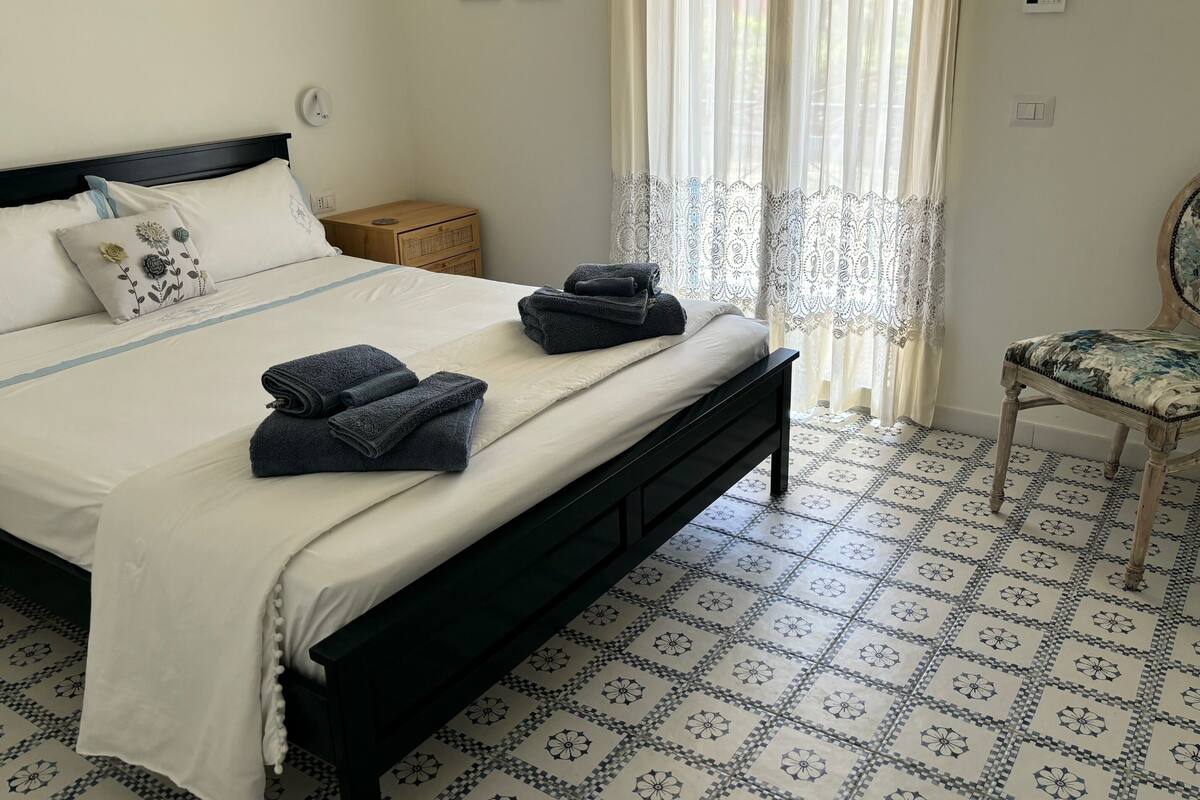 Un letto rifatto con asciugamani piegati, un comodino e una sedia con un cuscino floreale.