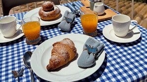 Ein Tisch mit karierter Tischdecke, ein Croissant, ein Gebäckstück, zwei Orangensaftgläser und eine Kaffeekanne auf einem Balkon mit Blick auf eine Landschaft mit Bäumen und blauem Himmel.