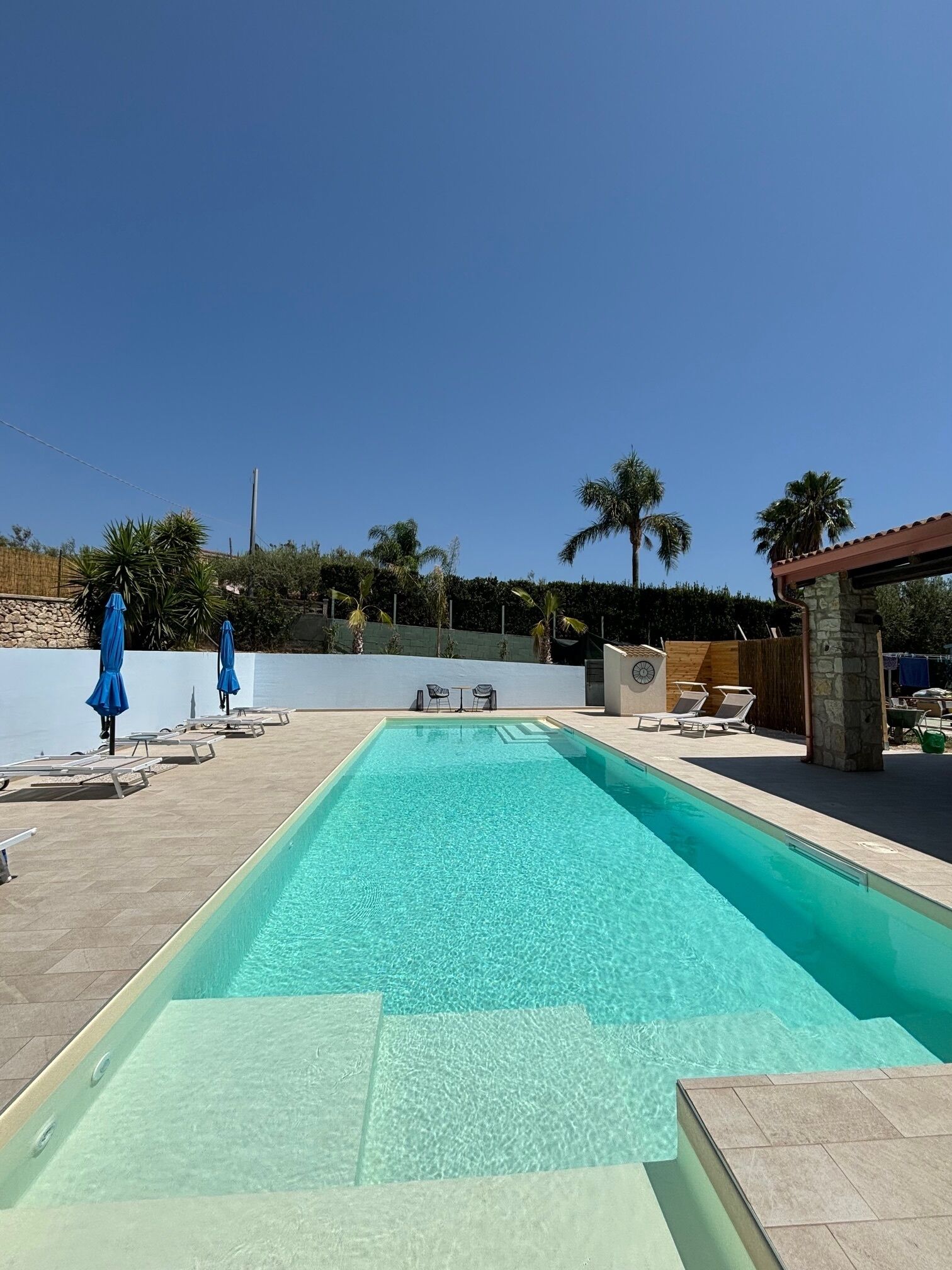 Una piscina rectangular con agua cristalina y azul, rodeada de sillones de descanso y sombrillas azules.