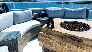 A cozy outdoor seating area with a wooden table, cushions, and binoculars, overlooking a body of water with mountains in the background.