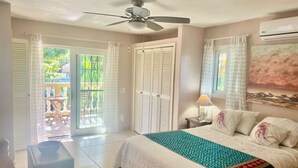A bedroom with a bed, a ceiling fan, a nightstand with a lamp, a window with curtains, and a door leading to a balcony. - Less than 2 minute walk to beach in half moon bay and main strip in west end (West End)