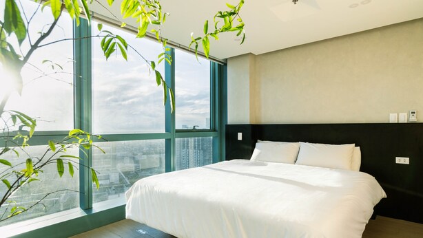 Quarto de hotel moderno com uma cama grande, vista da cidade por janelas do chão ao teto e uma planta no canto.