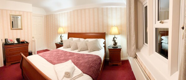 A hotel room with a large bed, two bedside tables with lamps, a dresser, a mirror, and a window with curtains. - Southlands B&B (Cheltenham)
