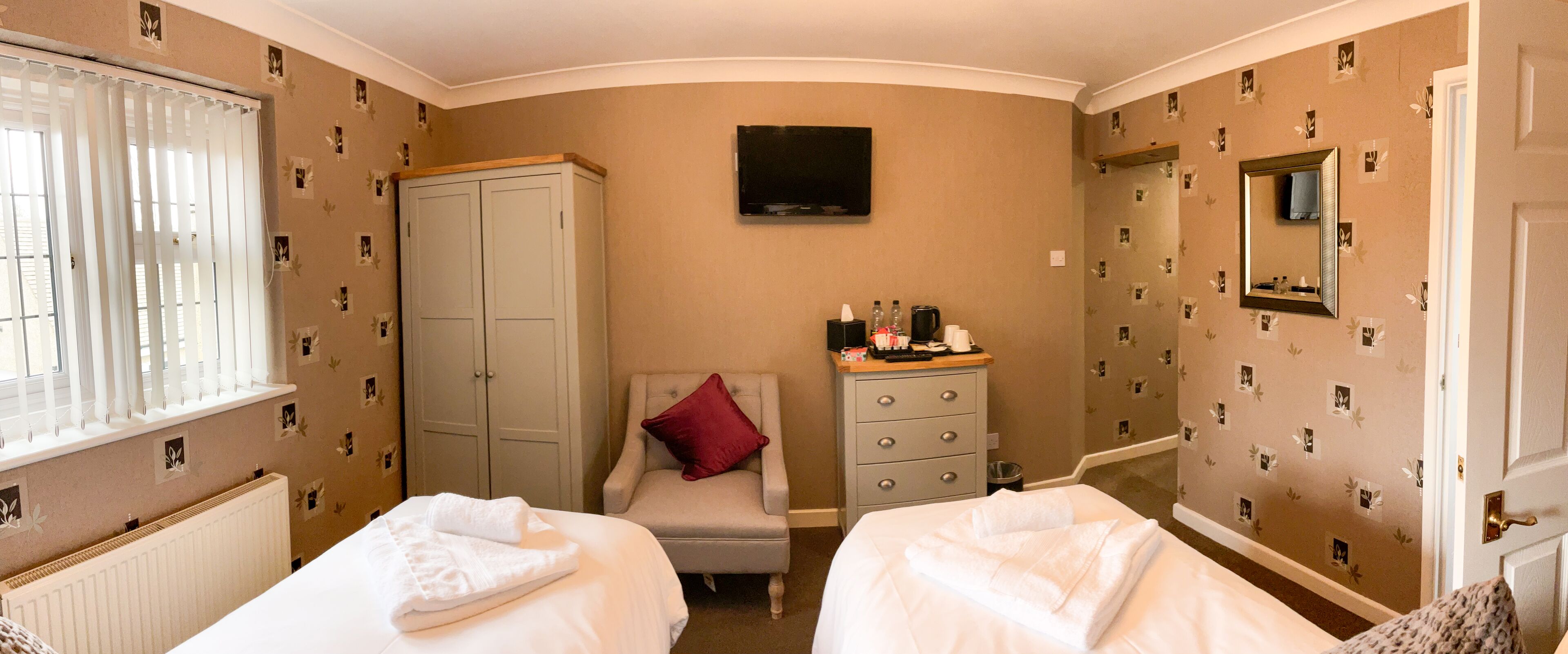 Ein Hotelzimmer mit zwei Betten, einem Kleiderschrank, einem Schreibtisch mit Toilettenartikeln, einem Fernseher und einem Spiegel.