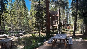 Une terrasse en bois avec des tables de pique-nique, située dans une zone boisée, avec un chalet en arrière-plan.