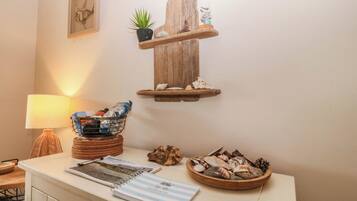 A room with a wooden shelf holding a plant and decorative items, a table with a lamp, a guest book, and a bowl of seashells.