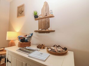 A room with a wooden shelf holding a plant and decorative items, a table with a lamp, a guest book, and a bowl of seashells. - Ty Ni (Dyffryn Ardudwy)