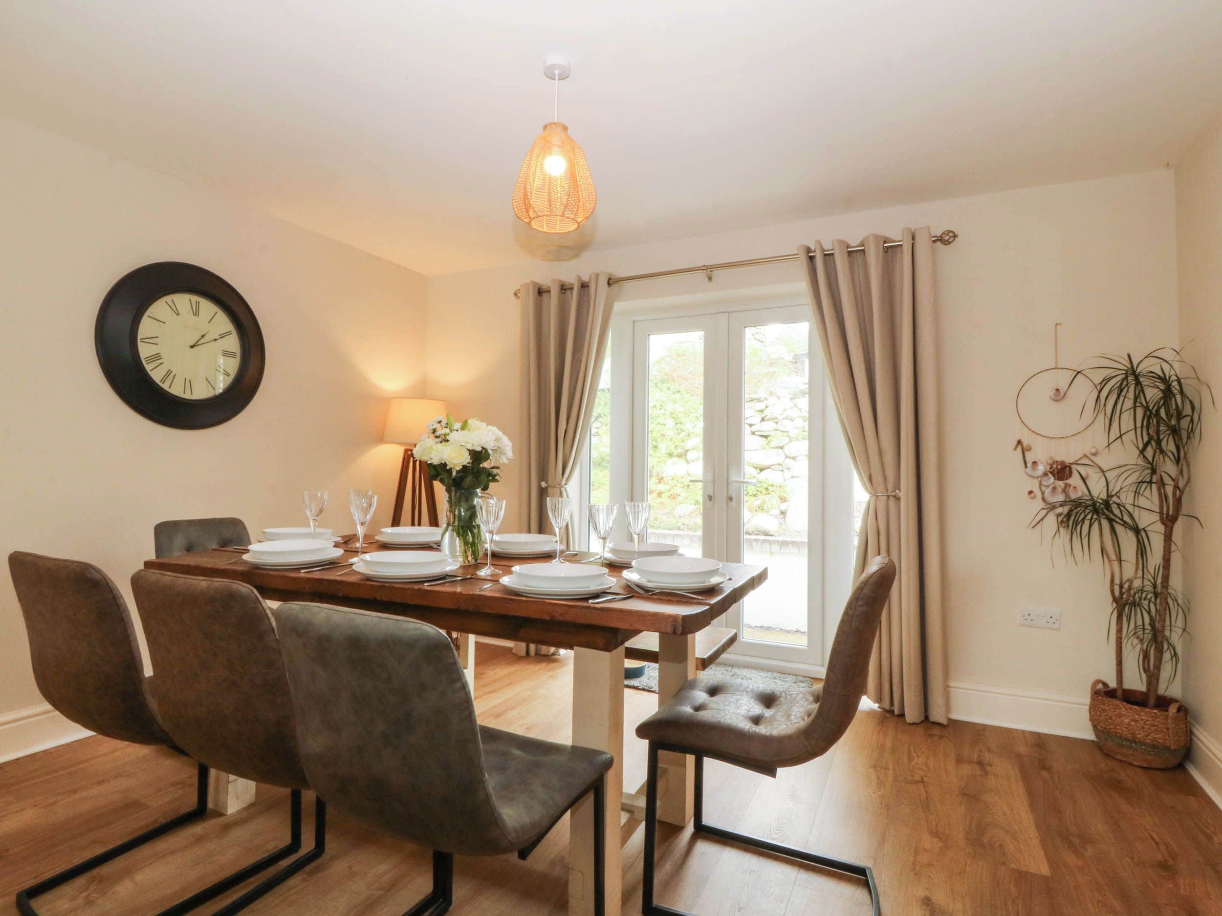 A dining area with a wooden table set for four, surrounded by upholstered chairs. A wall clock and a hanging light fixture are visible.