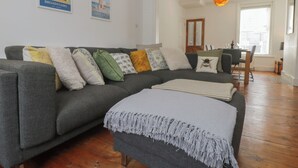 A living room with a grey sectional sofa, a small ottoman, and a dining area with a wooden table and chairs. There are two framed posters on the wall and a hanging pendant light. - 6 Sydney Road (Ramsgate)