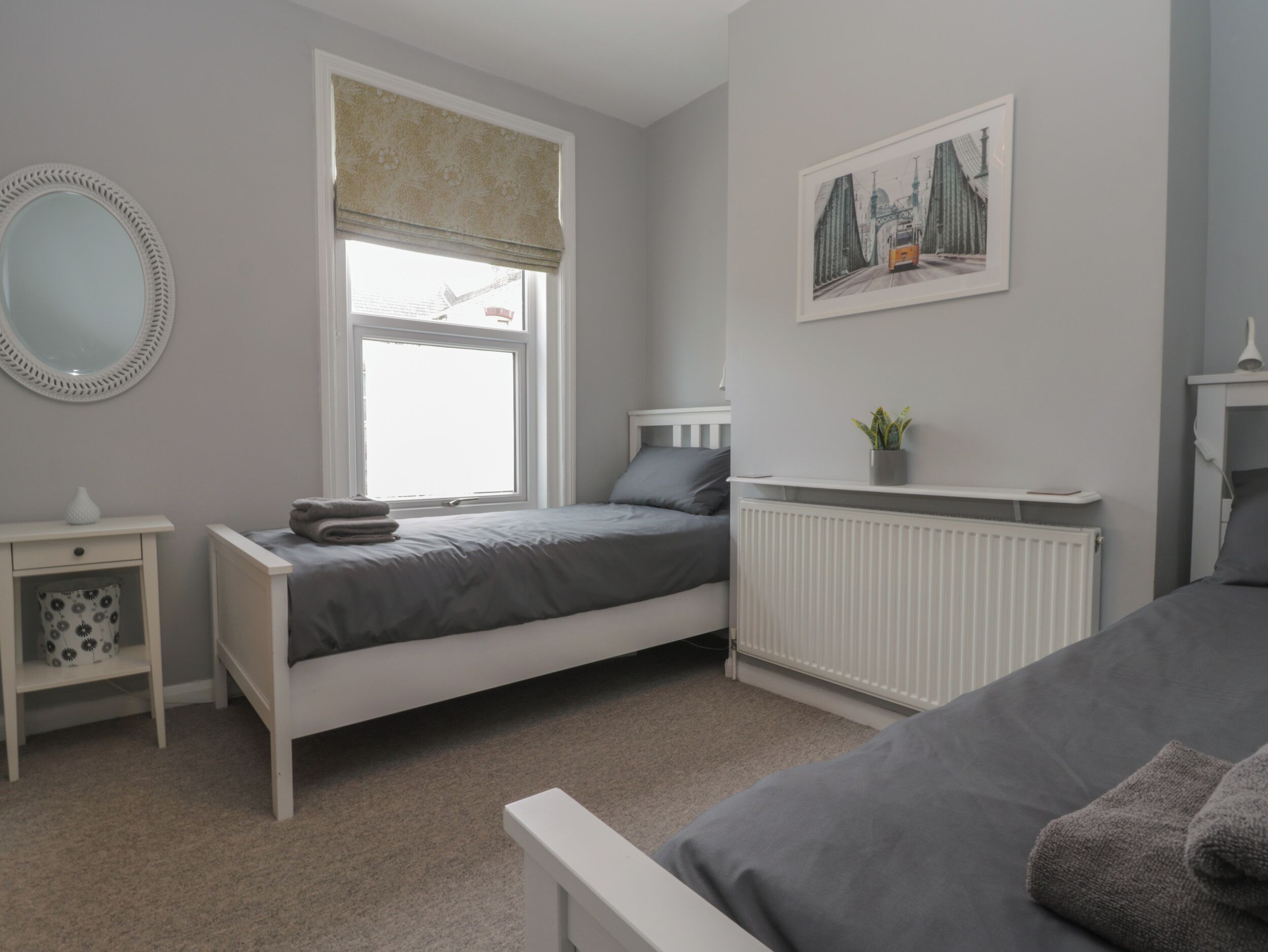 Une chambre avec un lit simple, une table de chevet, un radiateur, une fenêtre avec un rideau, un miroir et un tableau encadré au mur.