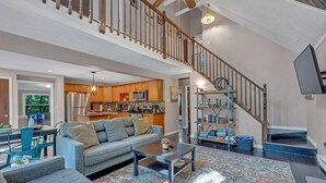 A spacious living area with a high ceiling, a staircase, and a kitchen in the background.