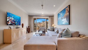 A modern living room with a flat-screen TV, a sofa set, a coffee table, and a view of the cityscape through large windows. - Beachfront, Premium Ocean View, 5 Star (Cabo San Lucas)