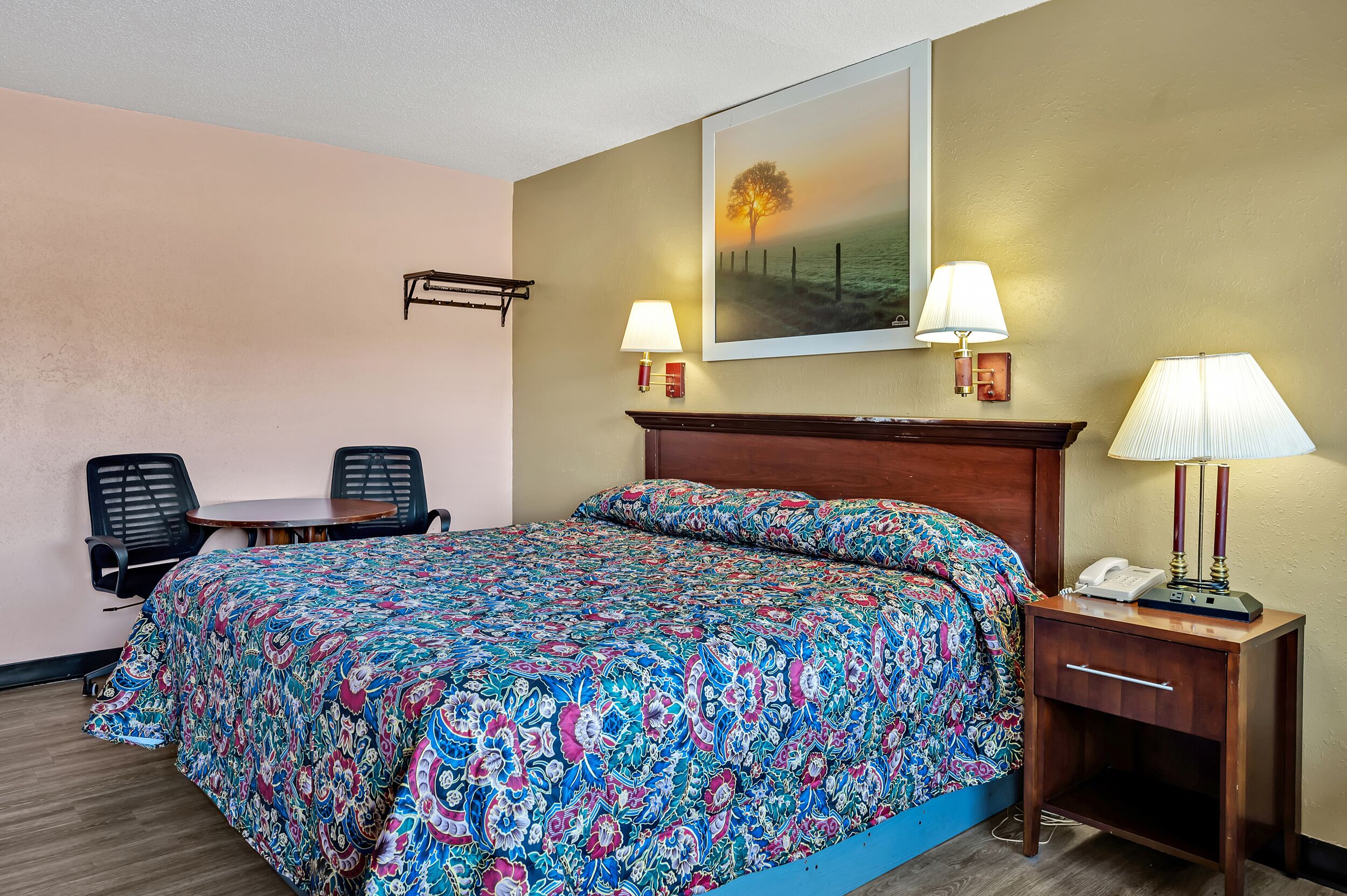 Quarto de hotel com uma cama grande, duas luminárias de cabeceira, uma mesinha com cadeiras e um quadro na parede.