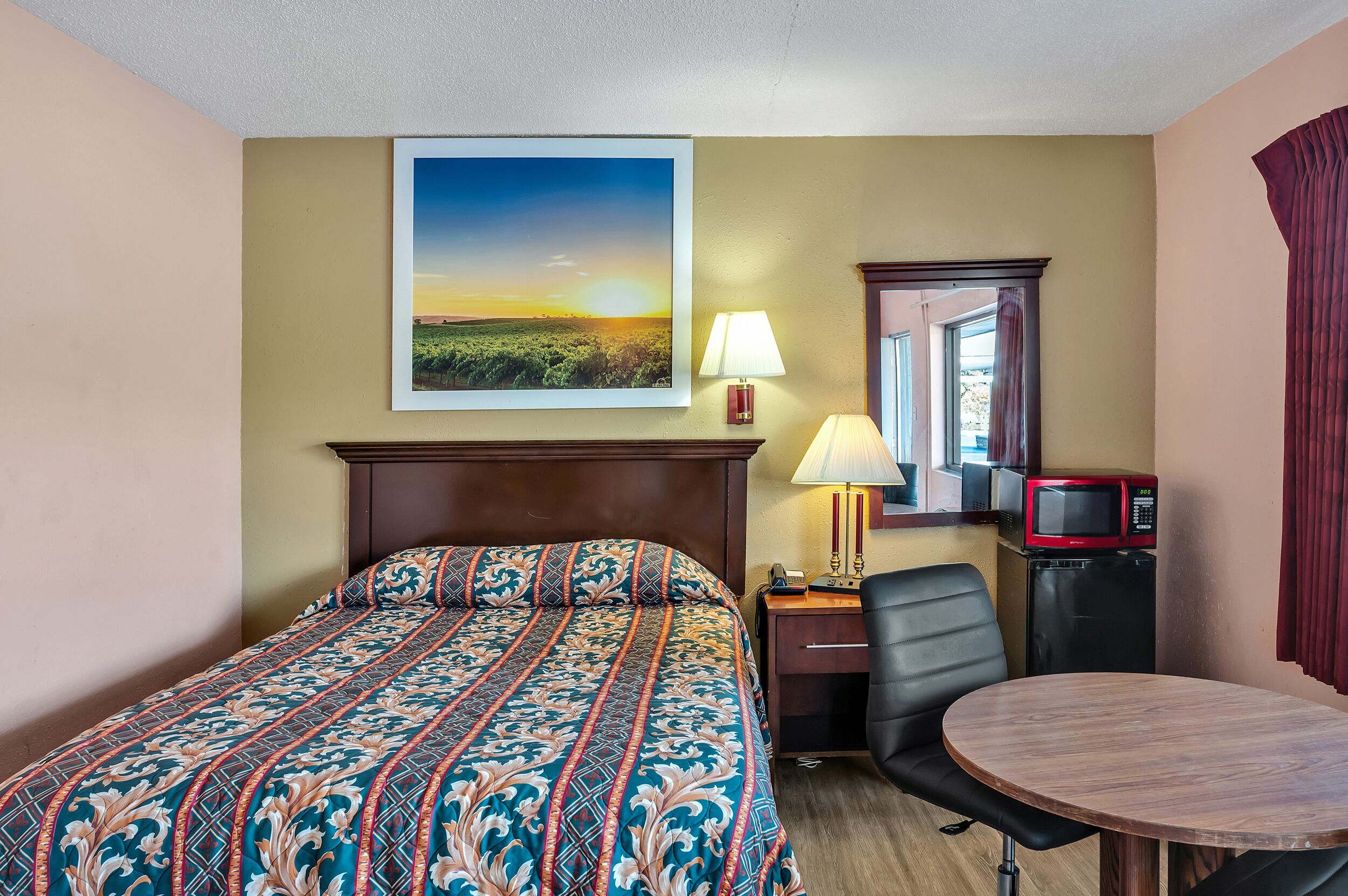 Habitación de hotel con cama, escritorio con lámpara, silla, mesa de comedor, microondas y un cuadro de un atardecer.