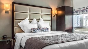 A hotel room with a large bed, a padded headboard, two wall-mounted lamps, and a window with a view of buildings.