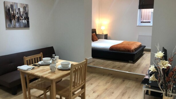 Une chambre d’hôtel moderne avec une table à manger en bois, un canapé, un lit recouvert d’une couverture marron et une table de chevet avec une lampe.