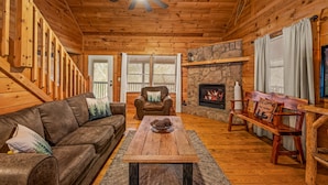 A cozy cabin interior with a wooden ceiling, a stone fireplace, a leather sofa, a wooden coffee table, and a bench.