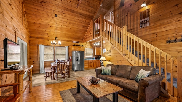 A wooden cabin interior with a living room, kitchen, and a staircase leading to an upper level.