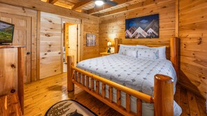 A wooden cabin bedroom with a bed, a ceiling fan, a painting, and a television on a wooden cabinet.