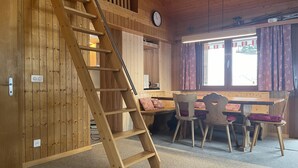 A wooden interior with a wooden staircase, a dining area with a table and chairs, and a window with curtains.