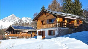 A wooden chalet with a balcony and solar panels, surrounded by snow and mountains.