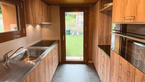 A narrow kitchen with wooden cabinets, stainless steel appliances, and a view of a garden through a door.