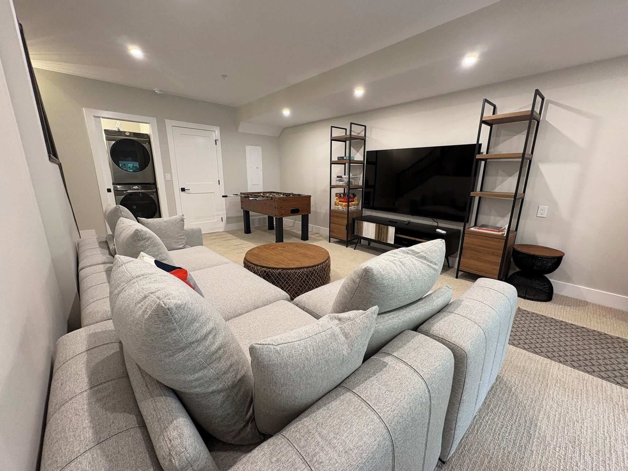 A modern living room with a sectional sofa, a foosball table, a flat-screen TV, and a bookshelf.