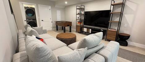 A modern living room with a sectional sofa, a foosball table, a flat-screen TV, and a bookshelf.
