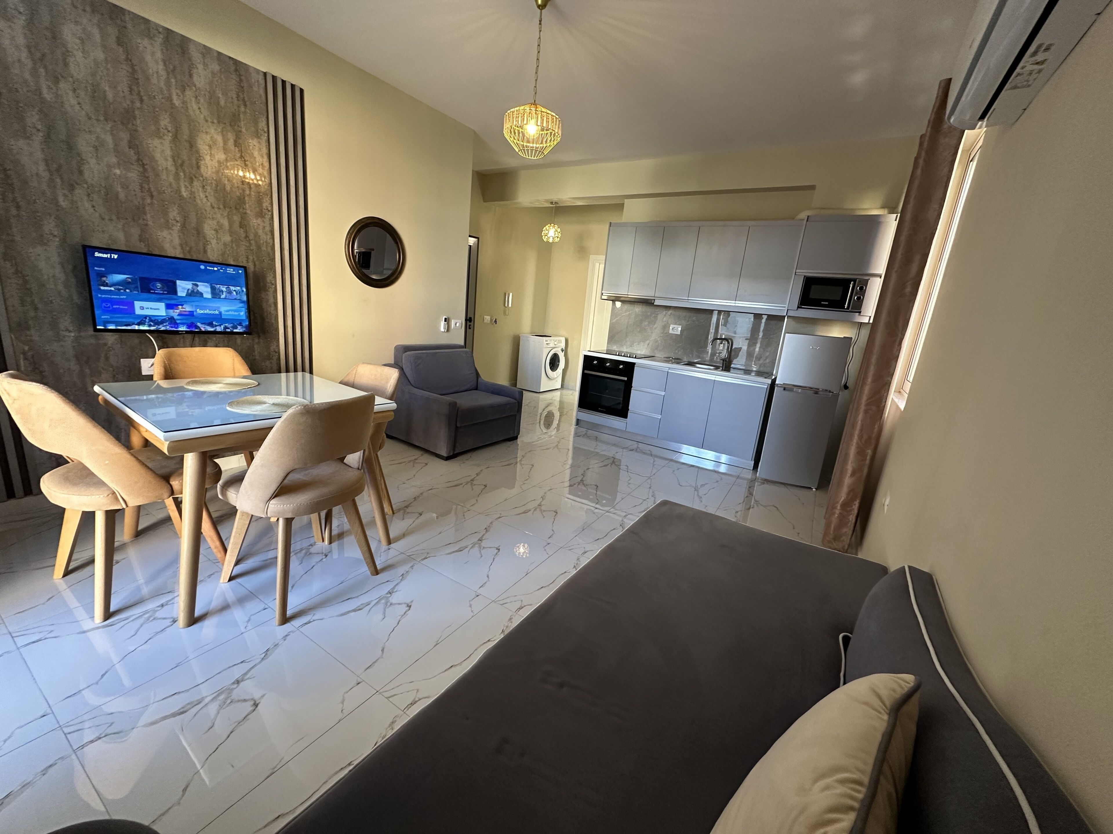 A modern living room with a glass dining table, comfortable seating, a grey sofa, and a kitchen area with stainless steel appliances.