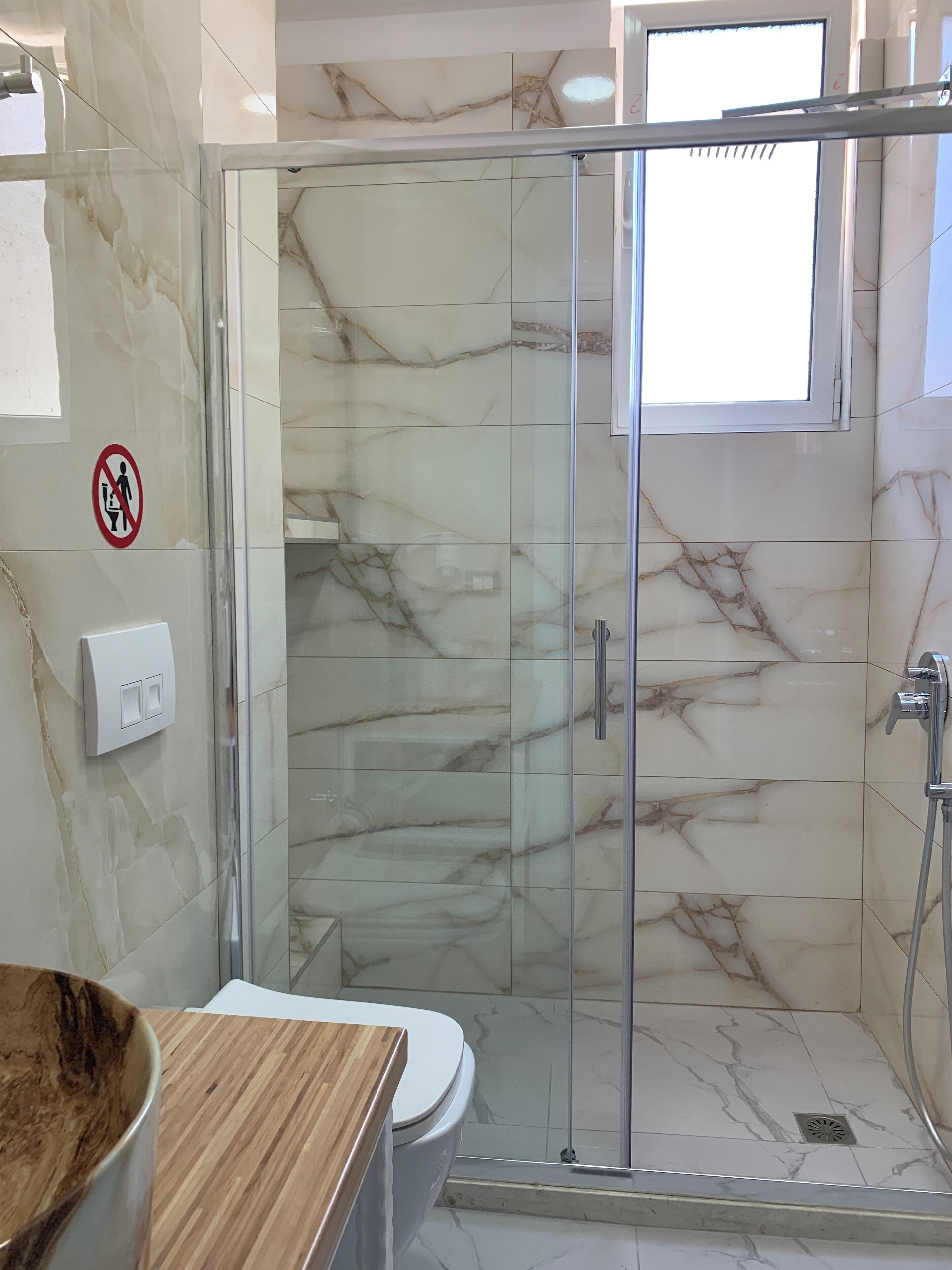 Un baño con ducha envuelta en vidrio, baldosas de mármol, un inodoro y encimera de madera.
