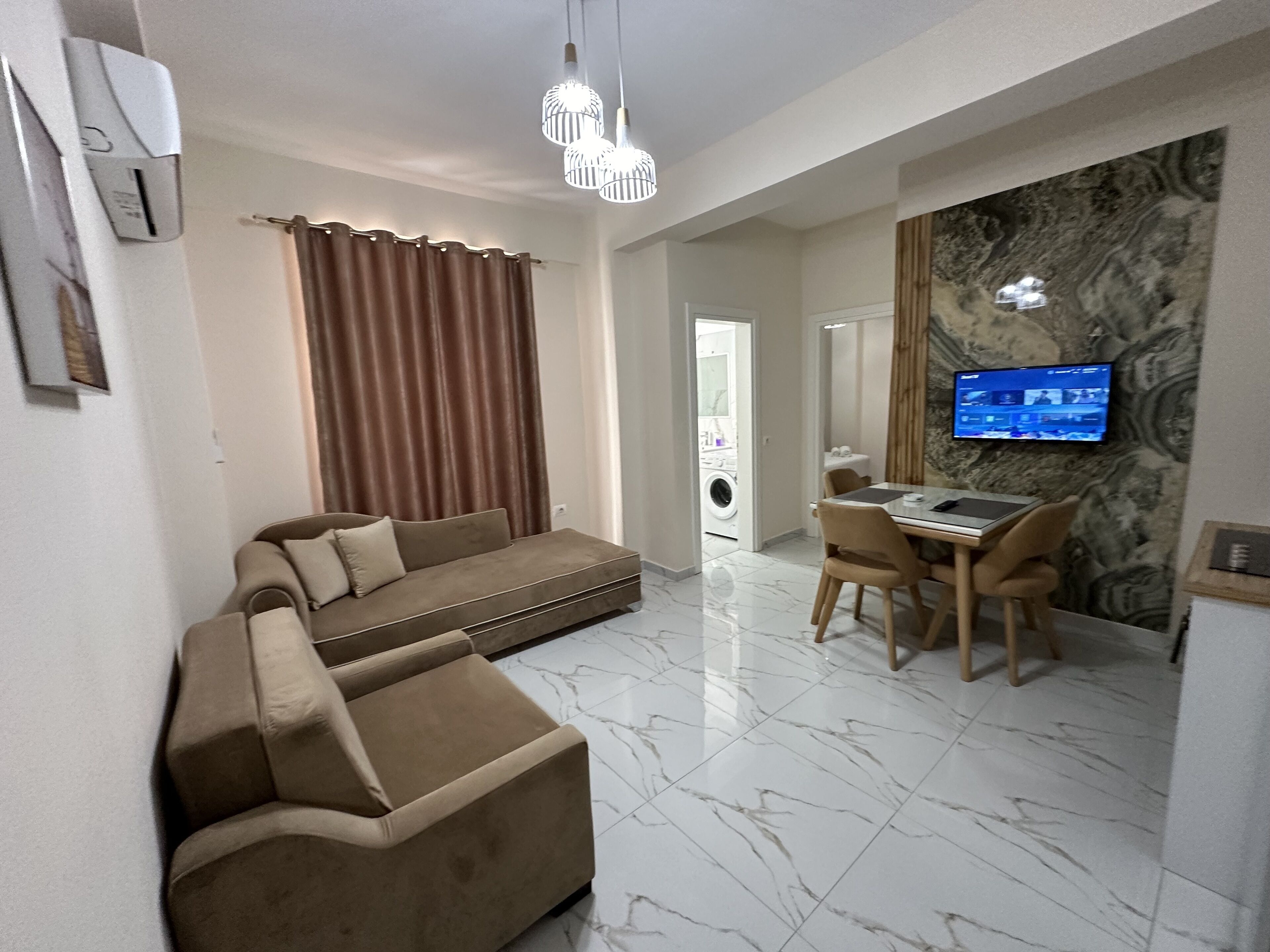 Una sala moderna con un sofá, una zona de comedor con mesa y sillas, un televisor montado en la pared y una lavadora al fondo.