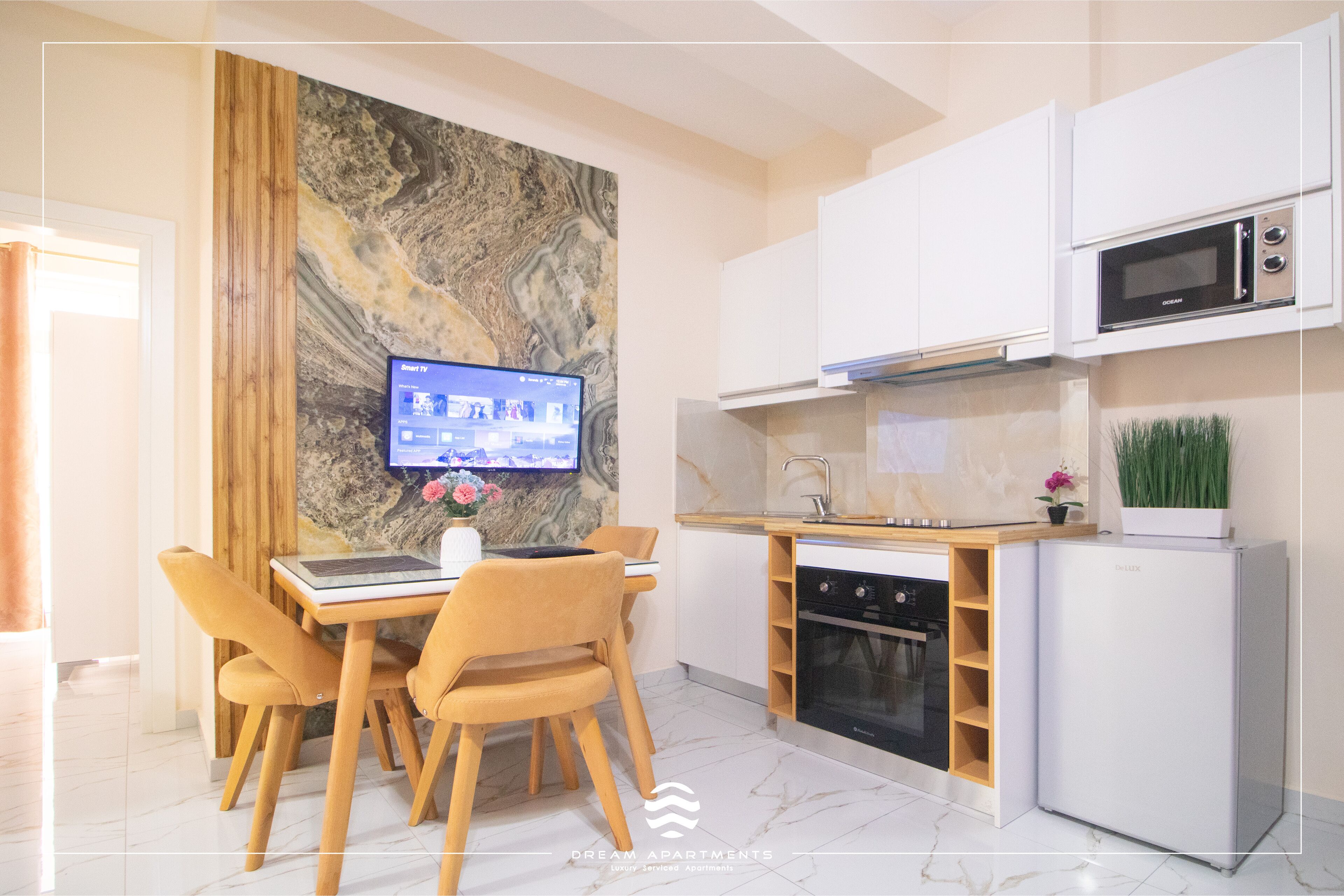 A modern kitchen with a built-in oven, microwave, and dishwasher. There is a dining area with a table and chairs, and a flat-screen TV mounted on the wall.