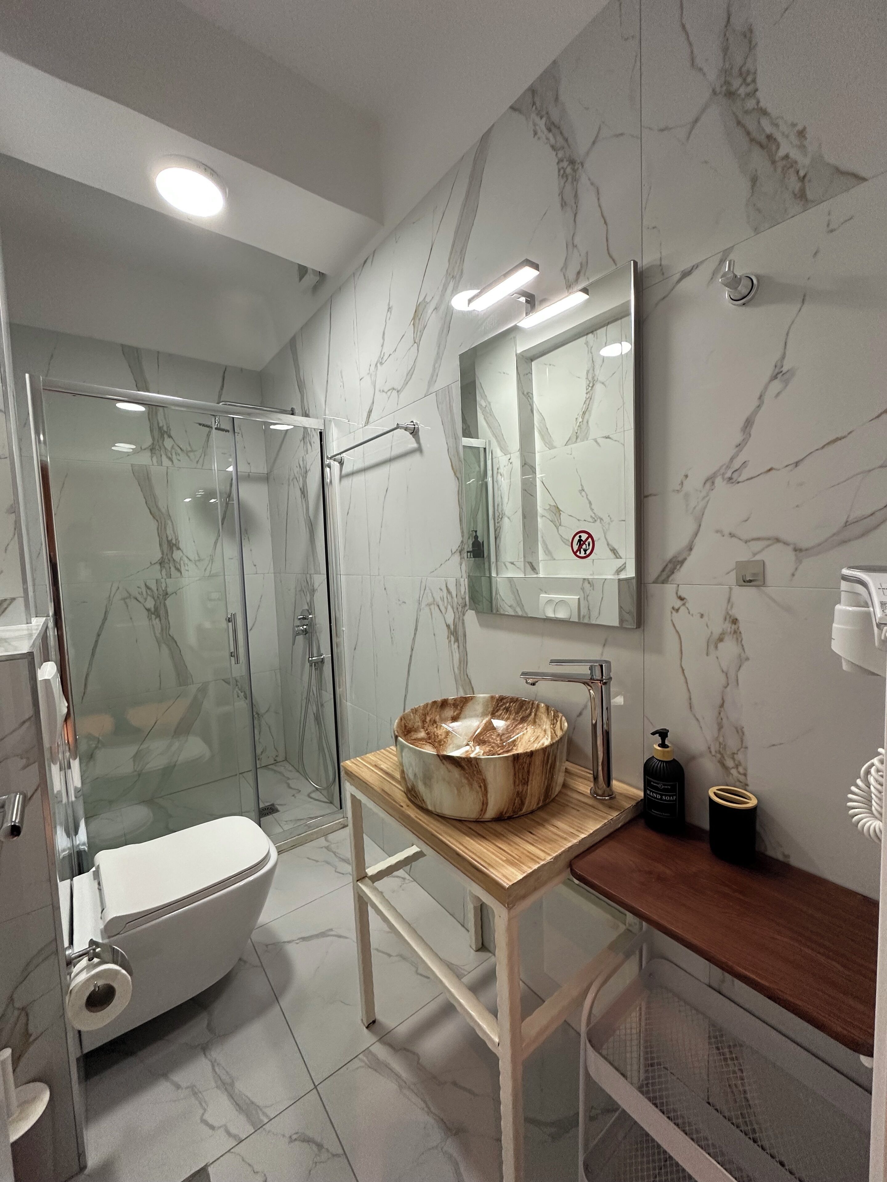 A modern bathroom with a glass-enclosed shower, a marble-tiled floor, a wooden vanity with a large basin, and a toilet.