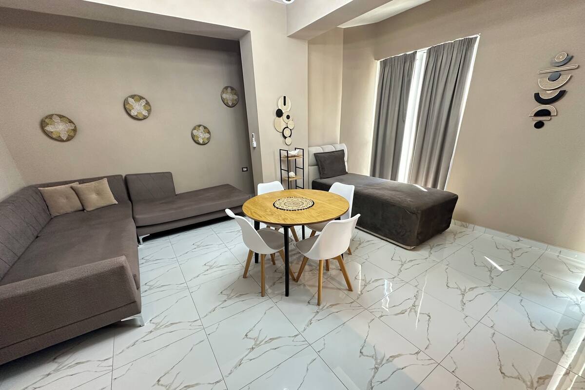 Un salon moderne avec un canapé d’angle, une table de salle à manger ronde entourée de chaises, un canapé gris et une grande fenêtre agrémentée de rideaux.