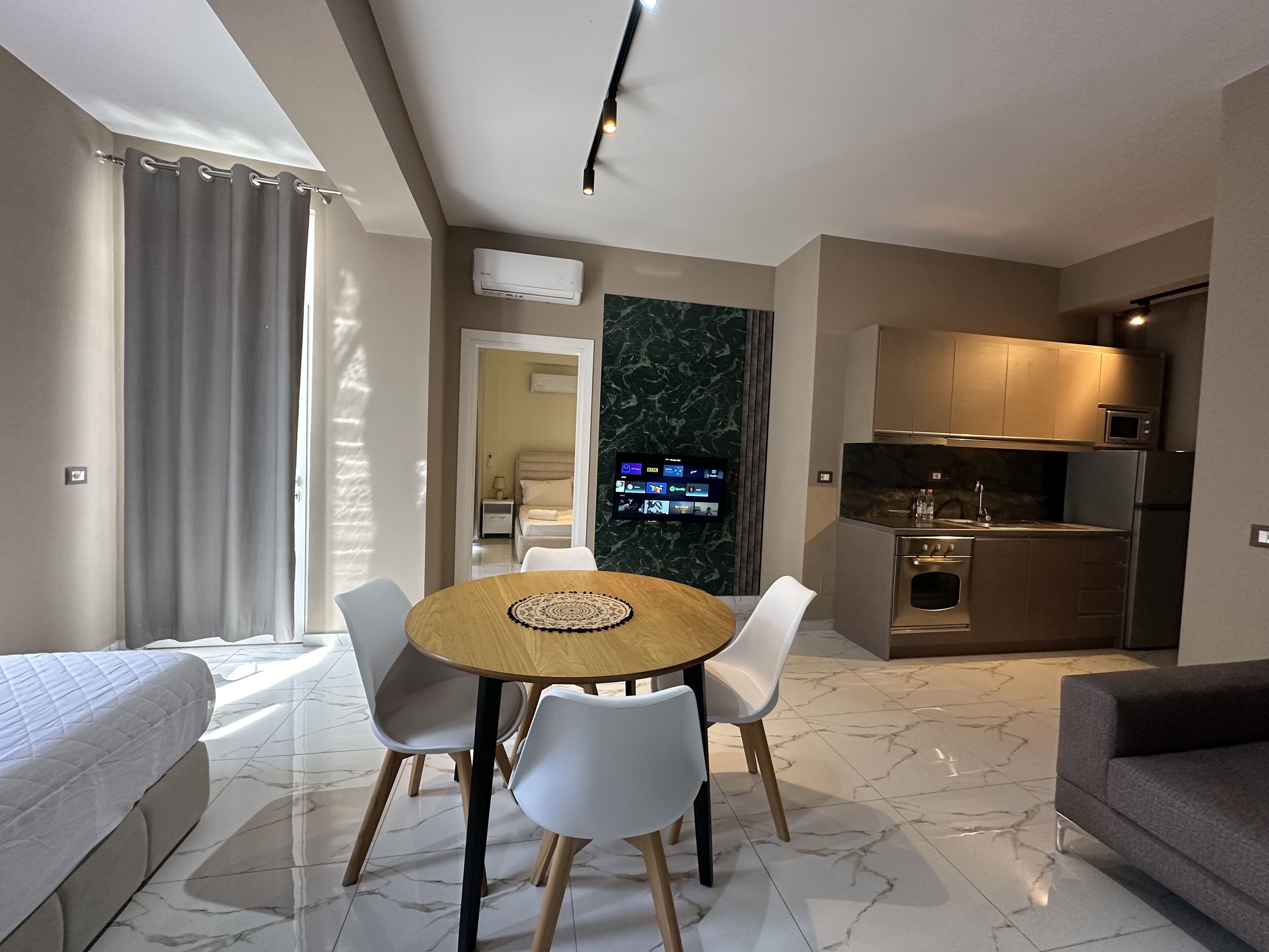 A modern hotel room with a dining area, kitchenette, and a TV mounted on the wall.