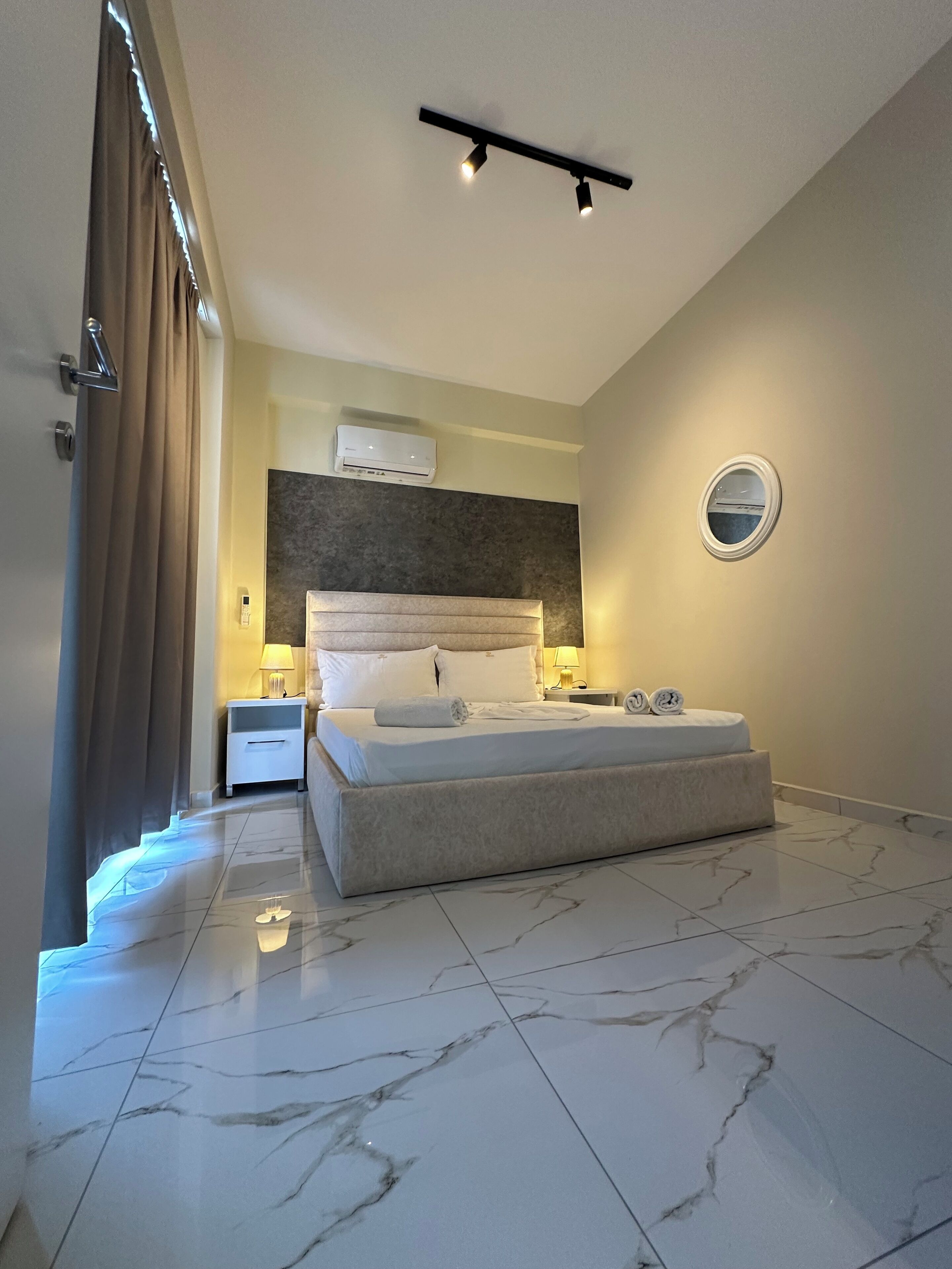 A modern bedroom with a large bed, a bedside table with a lamp, a wall-mounted air conditioner, and a round mirror.