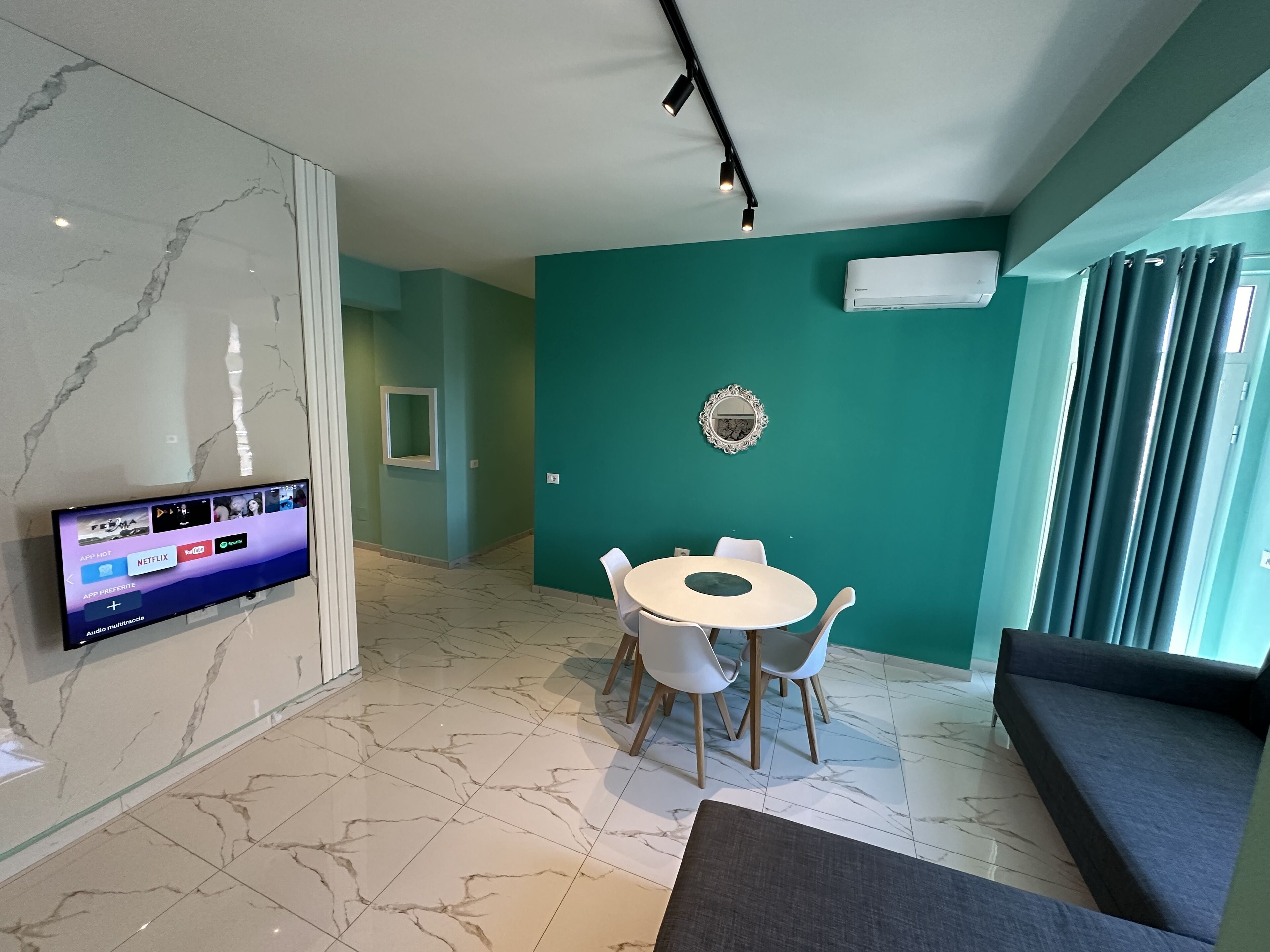 A modern living room with a marble floor, a flat-screen TV mounted on the wall, a round dining table with chairs, a sofa, and a large window with curtains.