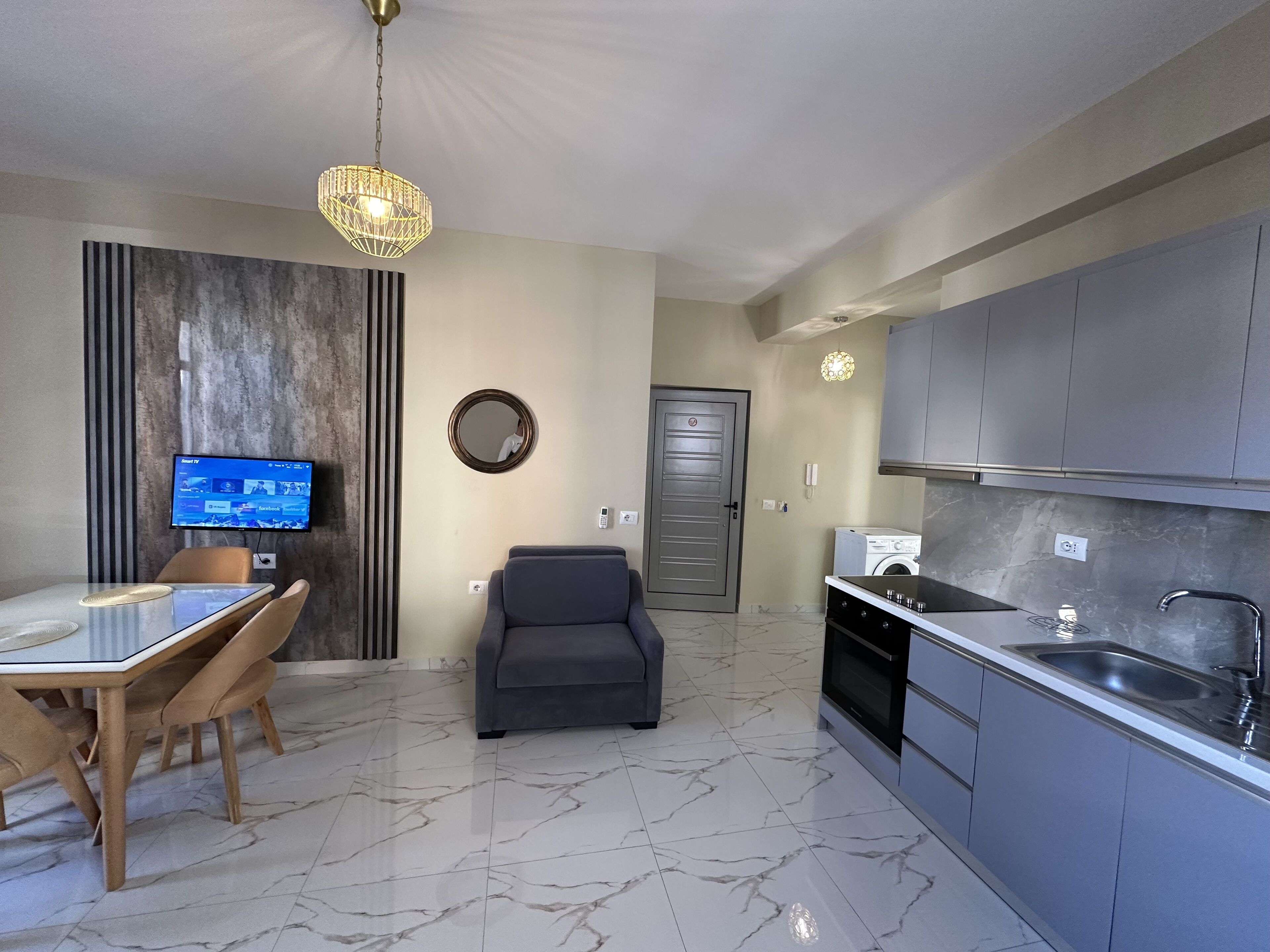 A modern kitchen with grey cabinets, a built-in oven, and a sink. There's a dining area with a table and chairs, a TV, and a grey sofa.