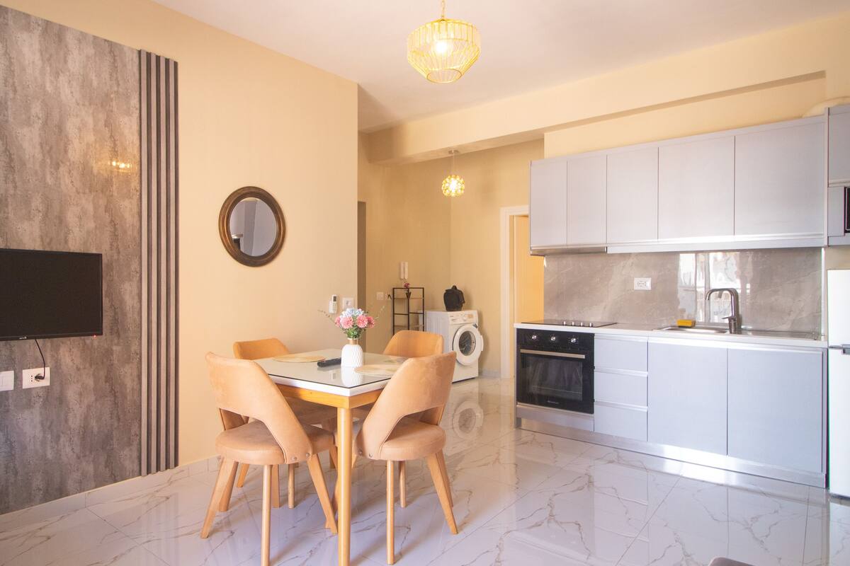 Une cuisine moderne avec des armoires blanches, un four encastré et un évier. On y trouve également un coin repas avec une table et des chaises, ainsi qu’un téléviseur fixé au mur.