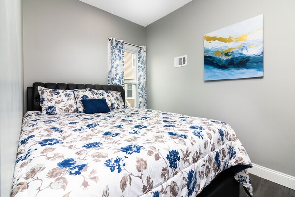 A bedroom with a bed, floral bedding, a window with curtains, and a painting on the wall. - Cozy 1 bedroom Apartment Sleeps 4 (Niagara Falls)
