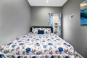 A bedroom with a bed, floral bedding, a nightstand, a window with curtains, and a wall-mounted air vent. - Cozy 1 bedroom Apartment Sleeps 4 (Niagara Falls)