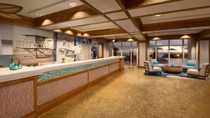 Un hall de hotel con mostrador de recepción, un ventanal con vistas a una zona de piscina y un área de descanso con sillas a rayas y una mesa de centro tejida.
